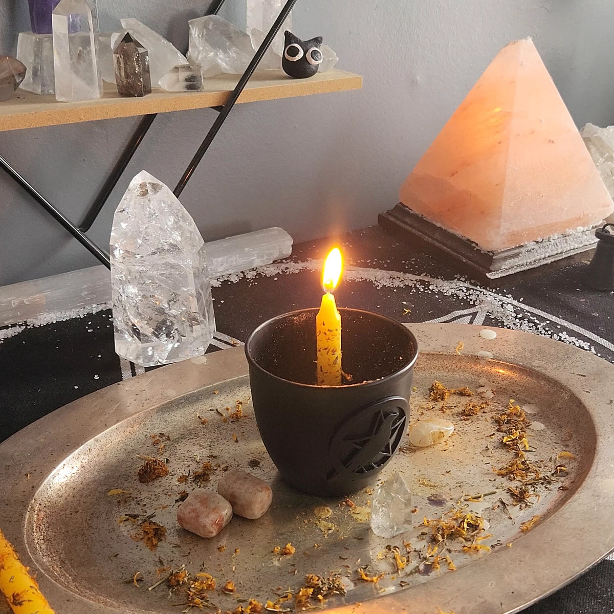 The Sunny Day Spell: a yellow candle dressed with herbs, lit in a black holder on an altar plate, with stones and herbs around it and a salt lamp behind