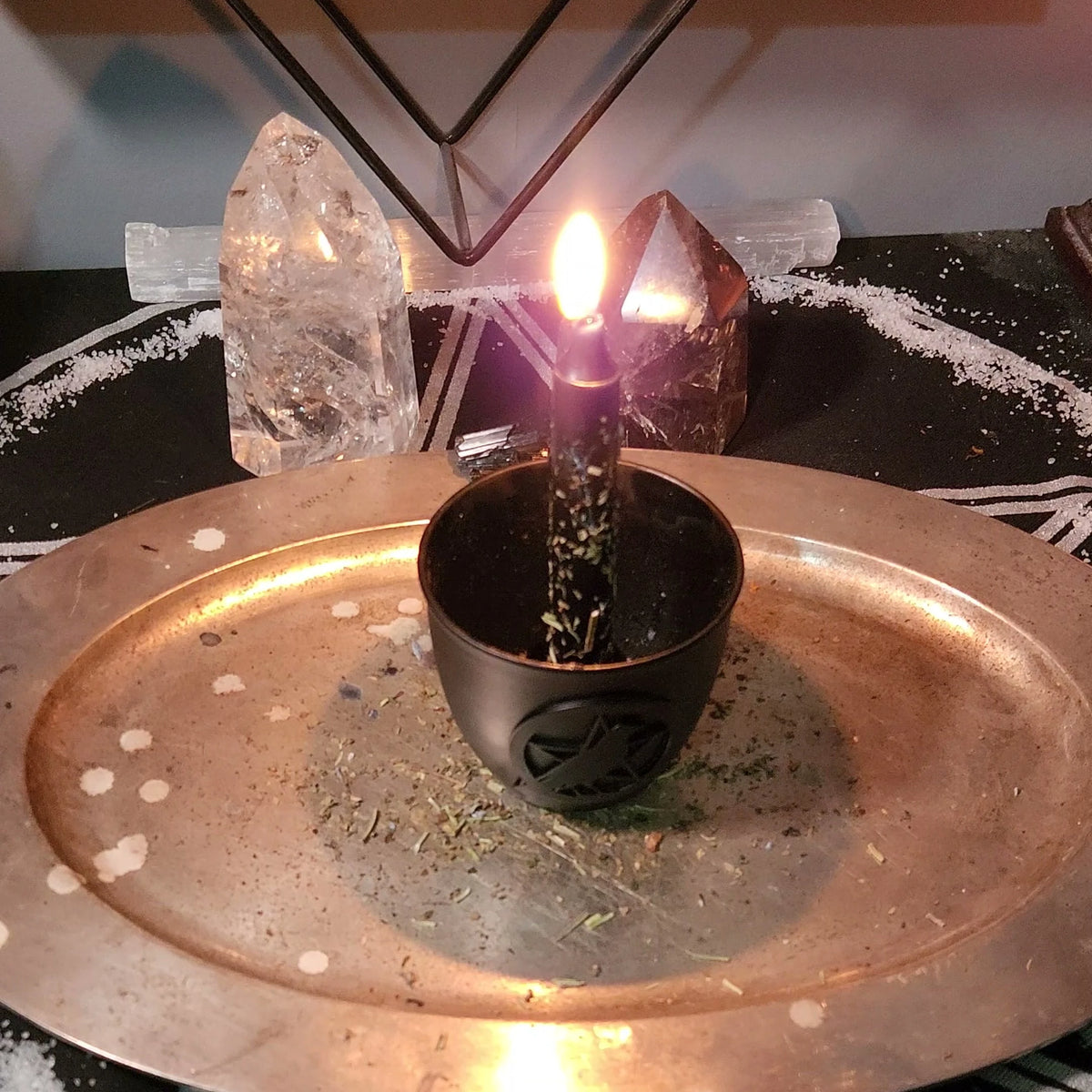 A black candle rolled in crumbled herbs, burning in a black holder on a round metal plate