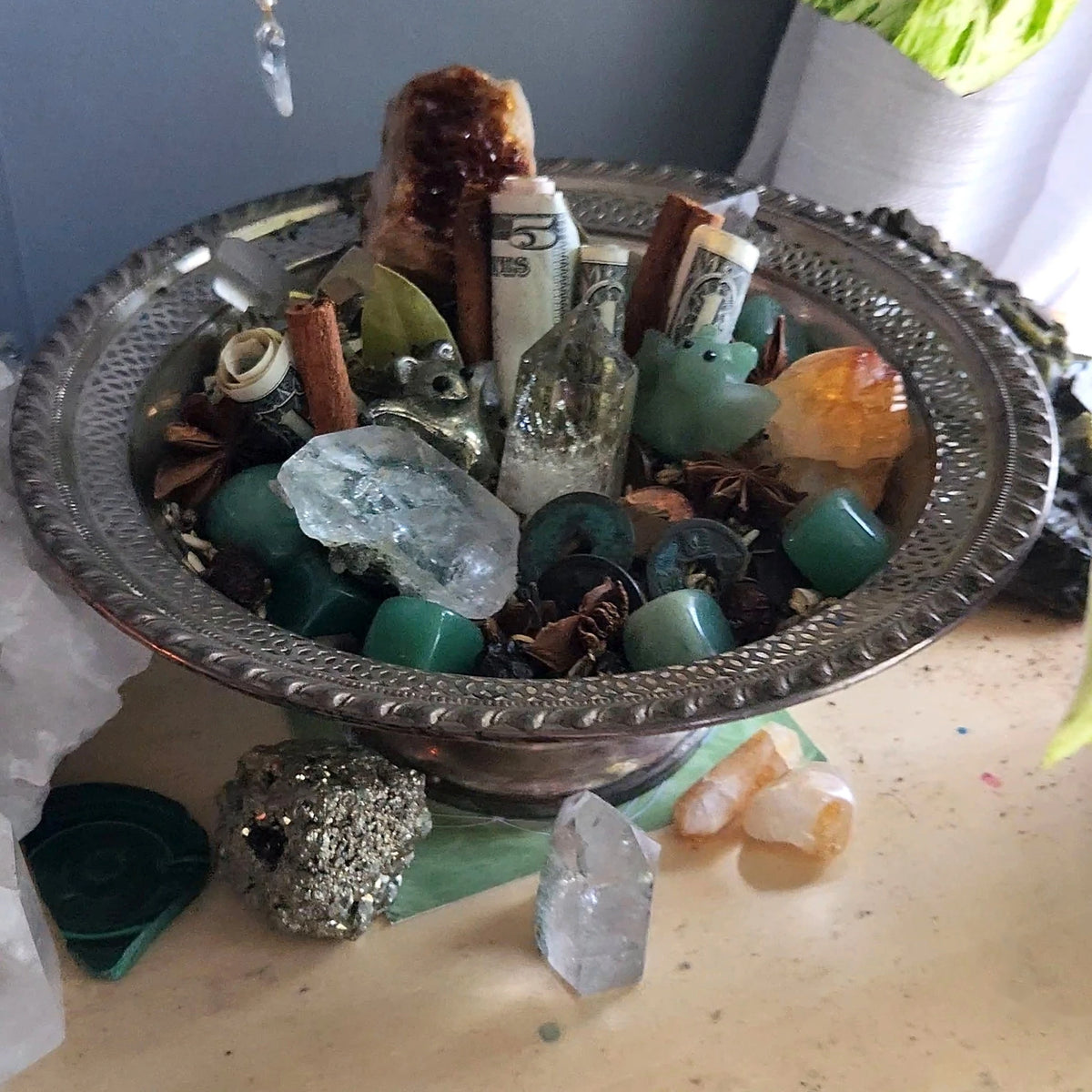 The Money Bowl: a silver bowl filled with rolled dollar bills, cinnamon sticks, star anise, coins, green aventurine, citrine and clear quartz, with pyrite beside it