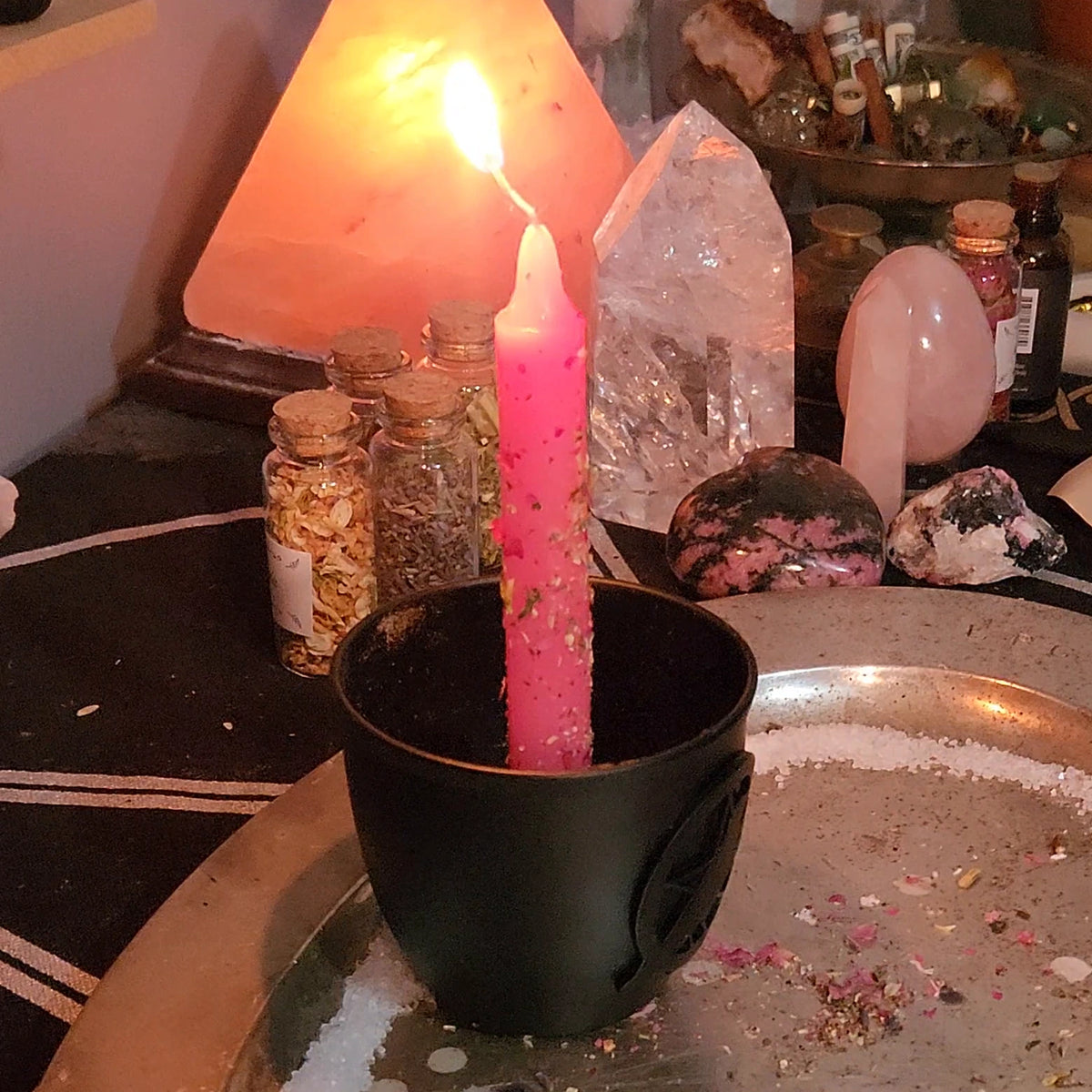 The Self-Love Candle: a pink candle dressed with herbs, lit in a black holder inside a ring of salt, with rose quartz and rhodonite behind it