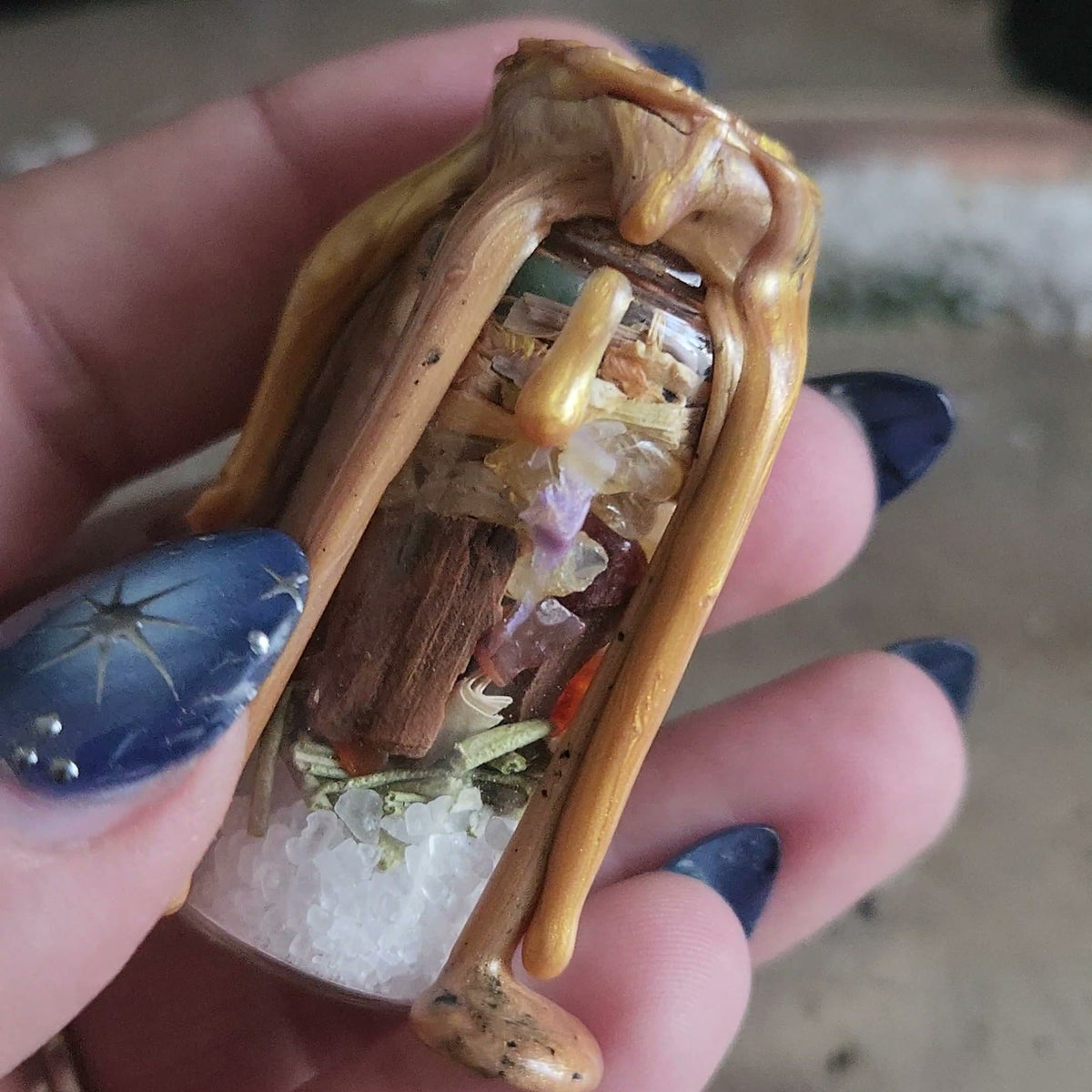 A small glass spell jar sealed with golden wax, held in a hand, with salt, herbs, a cinnamon stick and stones showing through the glass